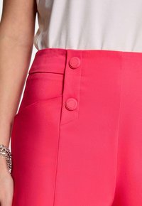 Pantalon fuchsia avec une texture lisse, deux boutons à la taille, poches latérales et une coupe ajustée.