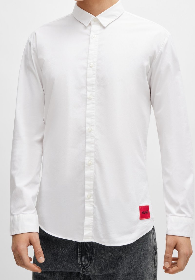 Man in een witte langemouwen overhemd met knopen, voorzien van een rood 'Hugo' label nabij de zoom, en zwarte gewassen jeans.