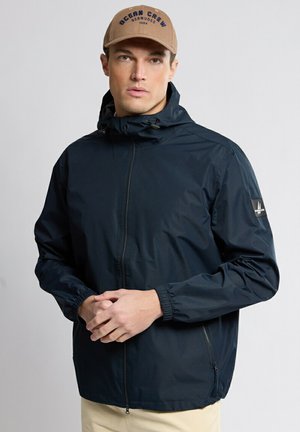 Homme portant une casquette de baseball marron clair avec un texte bleu marine et une veste zippée bleu marine avec des poignets élastiques, se tenant devant un fond clair uni.