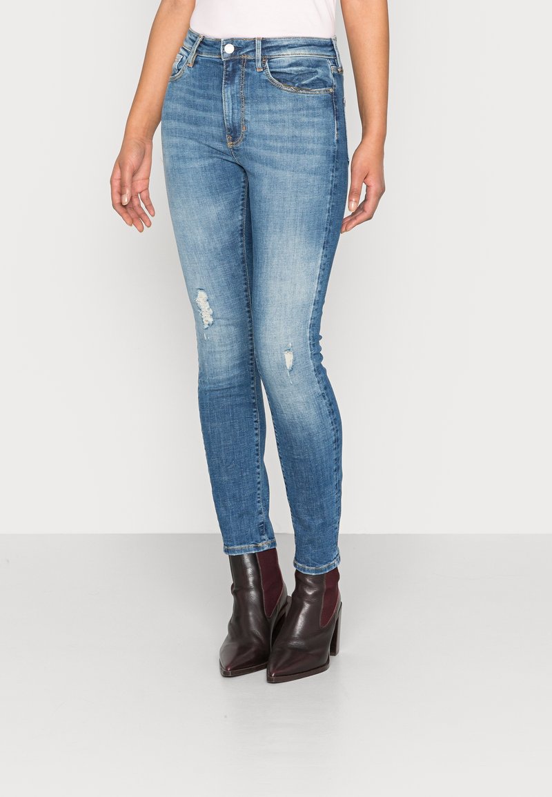Vaqueros ajustados de color azul con un acabado desgastado, que presentan detalles desgastados y un diseño clásico de cinco bolsillos, combinados con botas de tobillo burdeos.