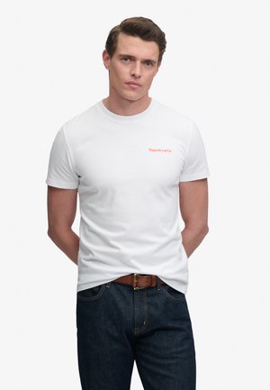 Mand iført en simpel hvid kortærmet T-shirt med lille rødt "Superdry&Co" logo, mørkeblå jeans og brunt bælte, stående med hænderne bag ryggen.
