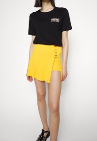 T-shirt en coton noir avec broderie florale, associée à une mini-jupe jaune côtelée avec une fente latérale et un détail en métal circulaire.