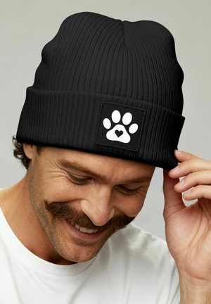 Man smiling and touching the brim of a black ribbed beanie with a white paw print and heart patch.