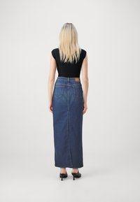 Jupe maxi en denim avec une taille ajustée, des poches arrière et une couture centrale. Portée avec un haut noir ajusté et des chaussures à talons. Couleur : bleu foncé.