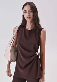 Top marrone senza maniche con design drappeggiato, caratterizzato da un dettaglio annodato e una texture liscia. Abbinato a una borsa rosa chiaro.