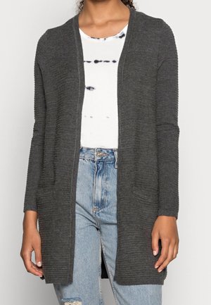 Kobieta ubrana w długi, ciemnoszary, otwarty kardigan na białą koszulkę z efektem tie-dye oraz jasnoniebieskie jeansy z przetarciami.