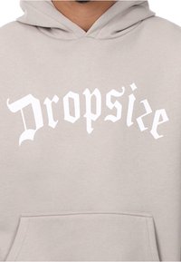 Hellgrauer Hoodie mit einem auffälligen weißen Drucklogo "Dropsie" in einer stilisierten Schriftart. Verfügt über eine Fronttasche und eine Kapuze mit Kordelzug.