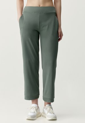 Person iført grønne cropped bukser med sidelommer og hvide sneakers, stående mod en ensfarvet lys baggrund.