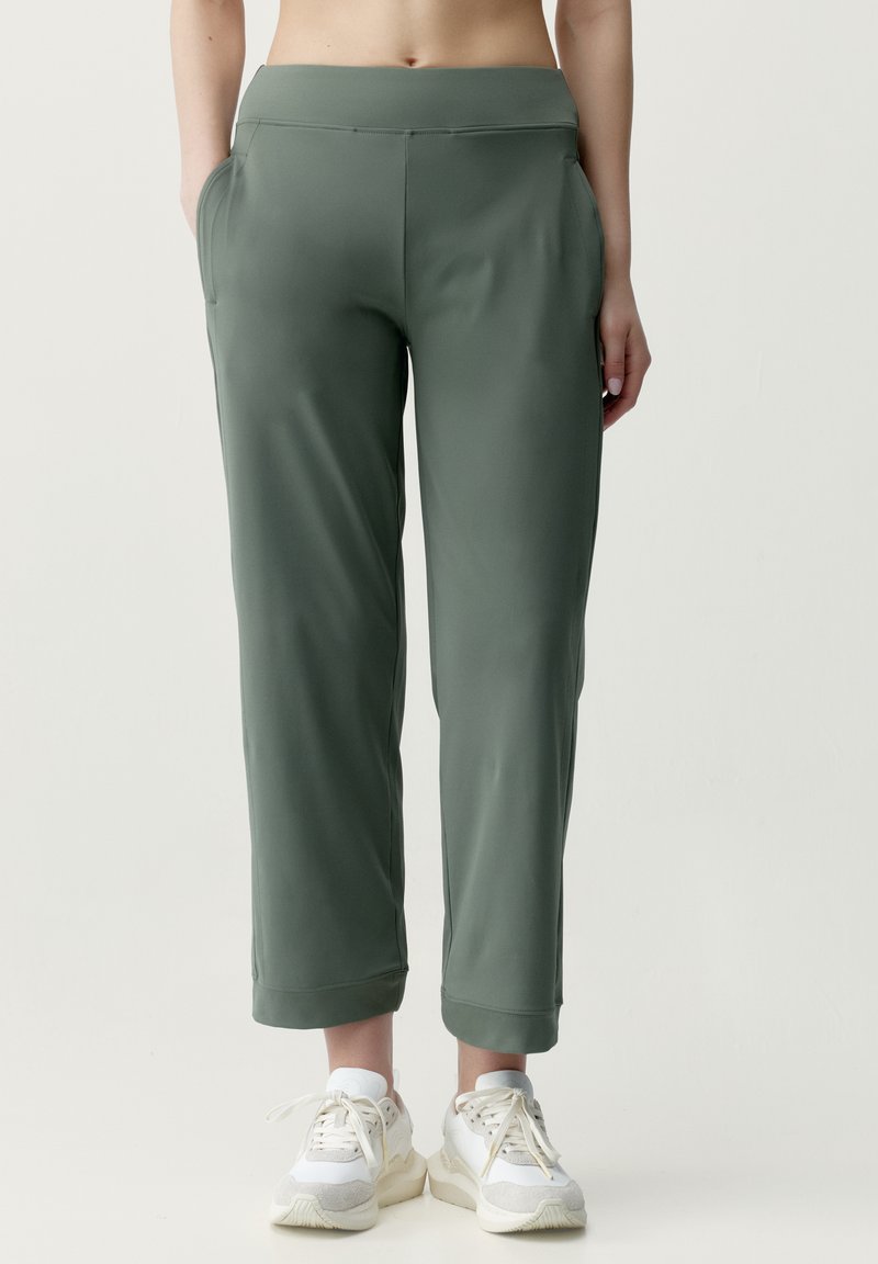 Personne portant un pantalon vert court à poches latérales et des baskets blanches, debout devant un fond clair uni.