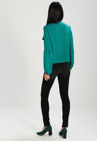 Kvinde står med ryggen til, iført en teal langærmet bluse, sorte stramme jeans og teal block-heeled sko foran en ensfarvet baggrund.