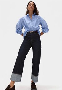 Modrá proužkovaná košile na knoflíky, tmavě denimové kalhoty s vysokým pasem a širokými nohavicemi se světle modrými ohrnutými lemy a tmavými botami. Jednoduchý design a střih.