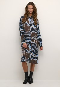 Vaatteena kuviollinen mekko, jossa on sininen, ruskea ja musta muotoilu; pitkät hihat, kaulus ja nappikiinnitys. Yhdistetty nilkkureiden kanssa.