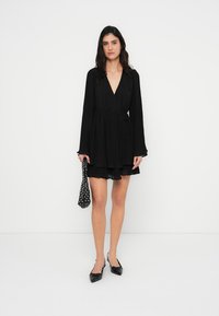 Μαύρο φόρεμα τύπου wrap με μακριά μανίκια, πλισέ υφή, λαιμόκοψη σε σχήμα V, στενή μέση, φούστα με φουσκωτή γραμμή, συνδυασμένο με αξεσουάρ που έχει πουά σχέδιο και επίπεδα παπούτσια.