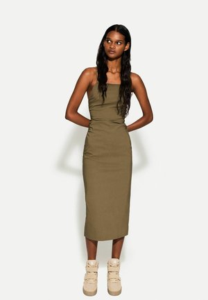 Abito midi senza maniche di colore verde oliva con corpetto aderente e dettagli arricciati, abbinato a scarpe alte beige con più fasce.