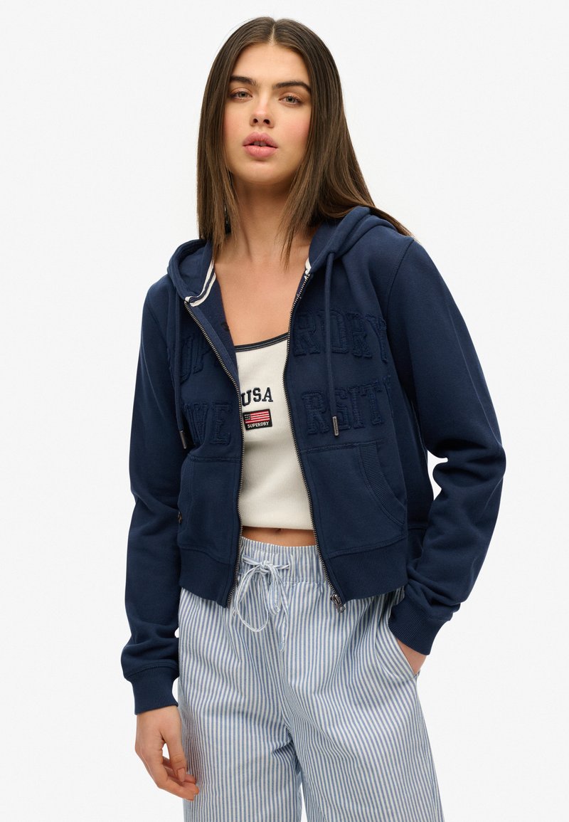 Veste à capuche zippée marine avec un design de texte en relief, tissu léger, deux poches avant, capuche avec cordon de serrage, portée par-dessus un débardeur blanc et un pantalon rayé.