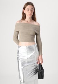 Yhdyksessä on beige hihaton ribbipaita, jossa on pitkät hihat, yhdistettynä kiiltävään hopeiseen istuvaan hameeseen. Musta käsilaukku pidetään kädessä.