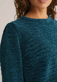 Teal stickad tröja med ett strukturerat vågmönster, rundad halsringning och lätt puffade ärmar. Mjuk, fluffig material med en avslappnad passform.
