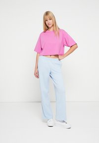 Ροζ κοντομάνικη cropped μπλούζα με κοντά μανίκια συνδυασμένη με ανοιχτό μπλε, άνετο παντελόνι φόρμας. Το μοντέλο φοράει λευκά αθλητικά παπούτσια. Απλό φόντο.