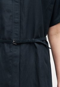 Κοντινό πλάνο σκούρου μπλε φορέματος με λεπτή ζώνη δεμένη στη μέση, που δείχνει την υφή του υφάσματος και μερικώς ορατό χέρι στη δεξιά πλευρά.