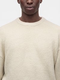 Homme portant un pull en maille côtelée de couleur crème, visible du cou à la poitrine sur un fond uni.