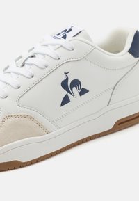 Zapatilla blanca fabricada con materiales de ante y cuero, que presenta un acento en azul marino, suela de goma texturizada y un diseño de logo en el costado.