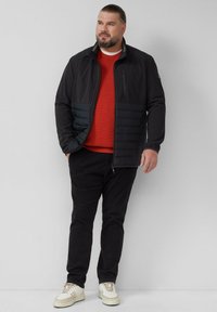 Giacca nera con pannello inferiore imbottito, tasche con zip e colletto a righe, indossata sopra un maglione rosso in maglia e pantaloni neri, abbinata a sneaker chiare.