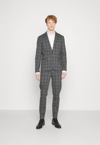 Grijze geruite pakken met een enkelvoudig jasje en slim-fit broek. Stof is gestructureerd en heeft een geknipt revers. Draagt een wit shirt en zwarte schoenen.