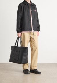 Czarny wiatrówka z zatrzaskami i elastycznymi mankietami, różowe logo, jasnoróżowa koszula pod spodem, beżowe spodnie, czarna torba na ramię z wytłoczonym logo.