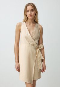 Beige wikkeljurk met een diepe V-hals, zonder mouwen en een strikdetail in de taille. Gemaakt van lichte stof met een gladde textuur.