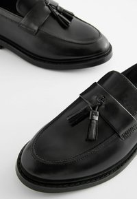 Mocassins en cuir noir avec une finition lisse, dotés d'une sangle décorative et de glands. Bout arrondi et détails de couture contrastés.
