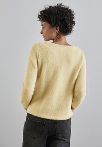 Maglione giallo, peloso, con una vestibilità rilassata, scollatura rotonda e maniche lunghe. Abbinato a jeans scuri, che mostrano una morbida trama.
