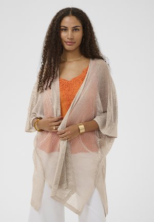 Donna con capelli ricci indossa uno scialle a rete beige sopra una maglia arancione di pizzo, adornata con bracciale, anello e collana dorati, in piedi davanti a uno sfondo bianco.