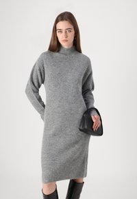 Graues gestricktes Sweaterkleid mit hohem Kragen, enganliegenden Ärmeln und gerippter Textur, kombiniert mit einer kleinen schwarzen Handtasche.