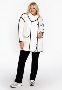Witte jas met zwarte bies, grote kraag en twee zakken aan de voorkant, gecombineerd met zwarte broek en zilveren sneakers. Gestructureerde stof.