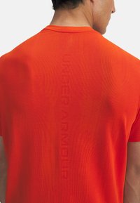 Rdeča kratka športna majica s teksturirano površino. Na hrbtni strani ima vertikalni logo "UNDERARMOUR" in standardno zasnovo okroglega ovratnika.