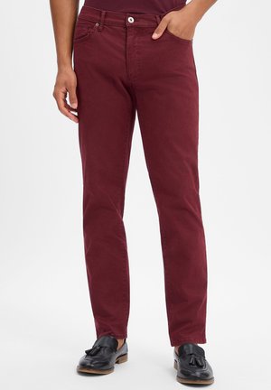 Persoon draagt slim-fit bordeauxrode broek met voorzakken, gecombineerd met zwarte loafers met franjes, staand tegen een witte achtergrond.
