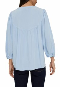 Blouse bleu clair avec des manches bouffantes, un col rond et des détails froncés au dos. Le tissu a une texture subtile. Poignets à boutons.