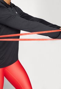 Schwarzes Langarmshirt mit Reißverschluss am Kragen, kombiniert mit engen roten Leggings. Ein rotes Widerstandsband wird zwischen den Händen gedehnt.