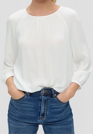 Persona con blusa blanca de manga larga con escote fruncido y vaqueros azules con las manos en los bolsillos, mostrada desde el cuello hasta las caderas.