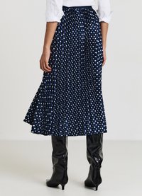 Jupe plissée bleu marine à pois blancs, en tissu brillant. La jupe tombe mi-mollet, associée à des bottes noires brillantes montant jusqu'aux genoux.