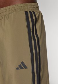 Oljen grønne treningsshorts med svarte striper; elastisk midjebånd; har et teksturert svart Adidas-logo på venstre side.