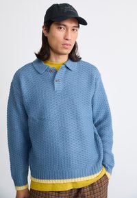 Ανοιχτό μπλε πλεχτό πουλόβερ με γιακά, δύο καφέ κουμπιά και ριμπ τελείωμα. Φορεμένο πάνω από ένα κίτρινο πουκάμισο και καφέ καρό παντελόνι.