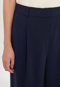 Pantalons larges bleu marine en tissu lisse, avec une taille cintrée et des plis. Gros plan montrant une partie d'une main reposant sur le côté.