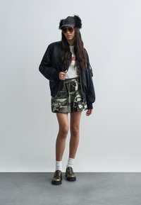 Czarna bomberka, biała koszulka z czerwonym nadrukiem, zielone szorty moro, puszysta czapka, duże okulary przeciwsłoneczne i masywne buty ze złotymi akcentami.