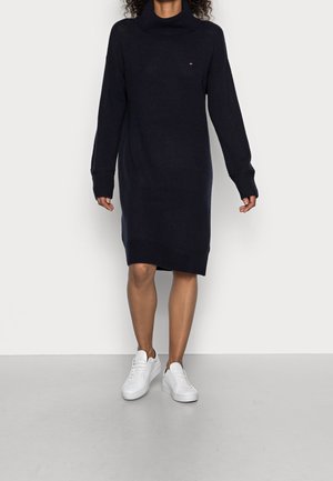 Frau trägt ein schwarzes langärmliges Pulloverkleid mit Rollkragen und weißen Sneakers, läuft auf einem grauen Boden vor einer weißen Wand.