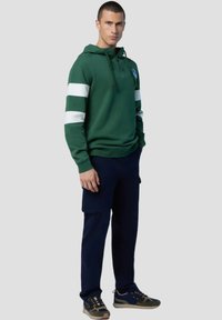 Felpa verde con cappuccio e strisce bianche sulle maniche, dotata di un collo zip e un patch. Abbinata a pantaloni cargo blu scuro e scarpe sportive.