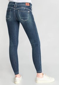 Jeans ajustados en un denim oscuro, con una cintura alta, diseño de cinco bolsillos, un ligero desgastado y costuras en contraste. Se llevan zapatillas blancas.