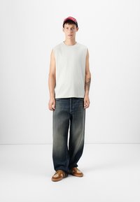 Lichtgrijs mouwloos shirt, donkerblauwe jeans en bruine sneakers. Rode pet met logo detail. Eenvoudige, casual stedelijke stijl.
