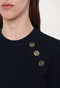 Gros plan d'une personne portant un pull côtelé bleu marine avec trois boutons dorés sur l'épaule, chacun gravé des lettres MK.
