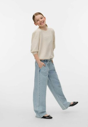 Lachend jong meisje met handen in de zakken, gekleed in een beige trui, losse lichtblauwe spijkerbroek en zwarte balletpantoffels, staand op één been.